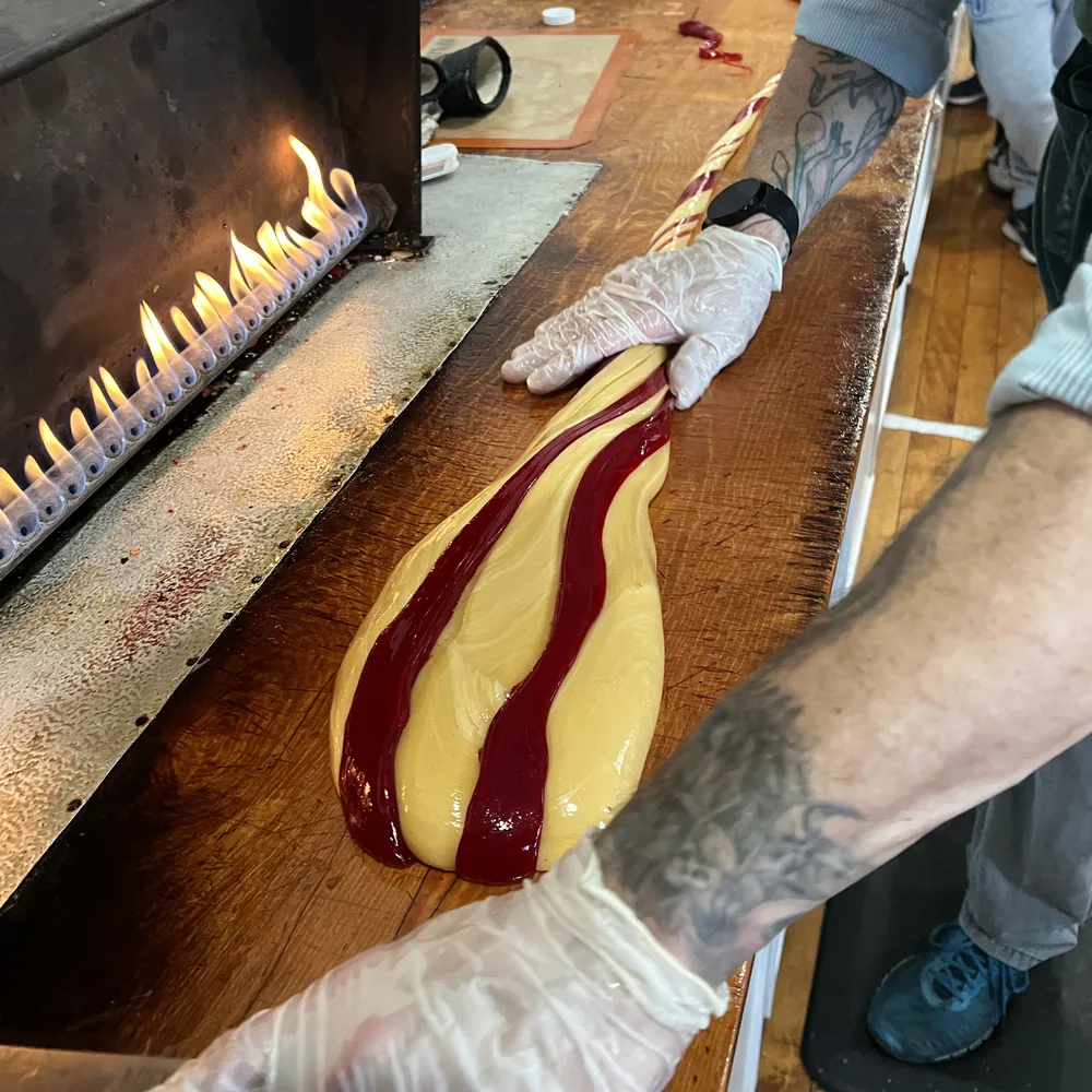 Candy makers pulling a warm red-and-gold striped candy cane batch across the table next to the flame